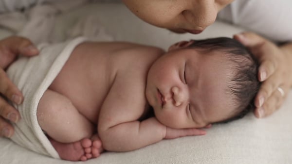 Sleeping newborn swaddled in cream wrap, mother leaning in to kiss the top of the head