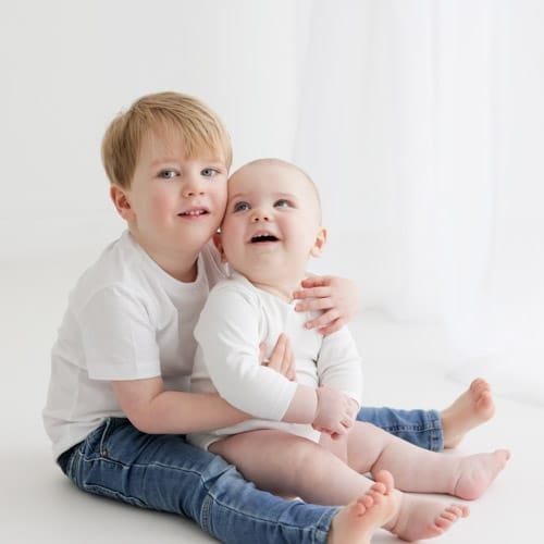An older brother sitting on the floor holding his baby sibling, both laughing