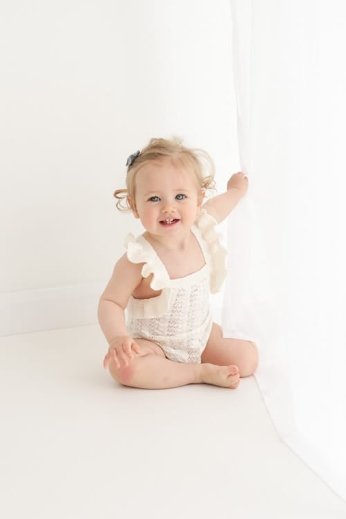 Baby in a cream romper sitting by a white curtain, holding it with one hand
