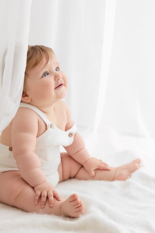 Baby sitting on a white blanket beside a voile curtain, looking upward