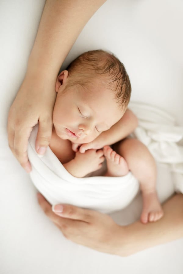 Newborn curled in a white wrap, held in a parent's hands, seen from above