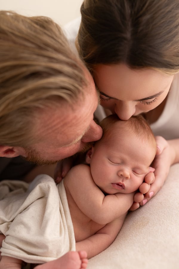 Two parents kissing, their sleeping newborn wrapped on the blanket below them