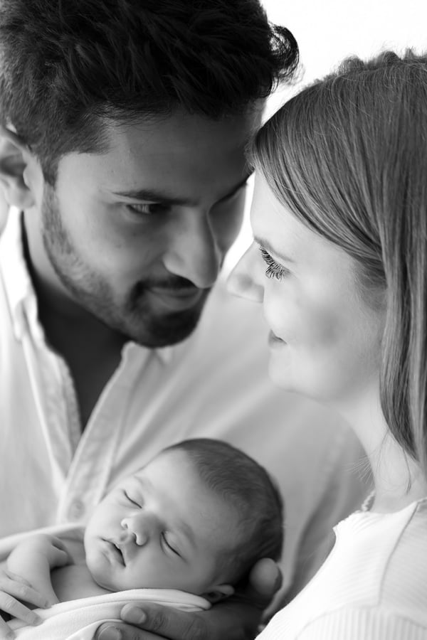 Both parents holding their swaddled newborn, the father in profile, in black and white