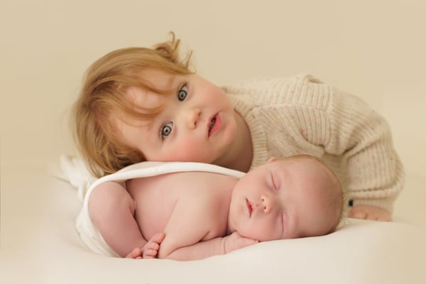 A toddler in a cream knitted jumper lying behind the sleeping newborn, looking at the camera