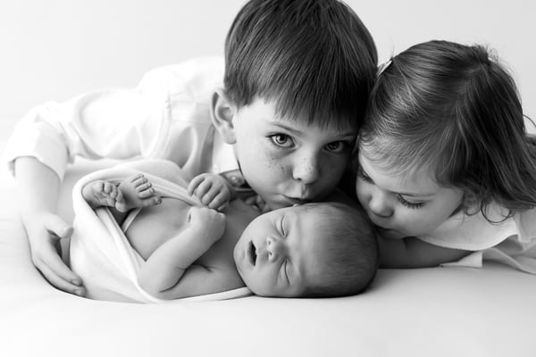 Two older siblings leaning in from either side to kiss their sleeping newborn sibling