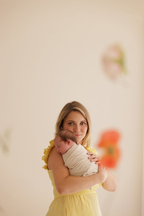 A mother in a yellow dress holding her swaddled newborn, flowers behind them
