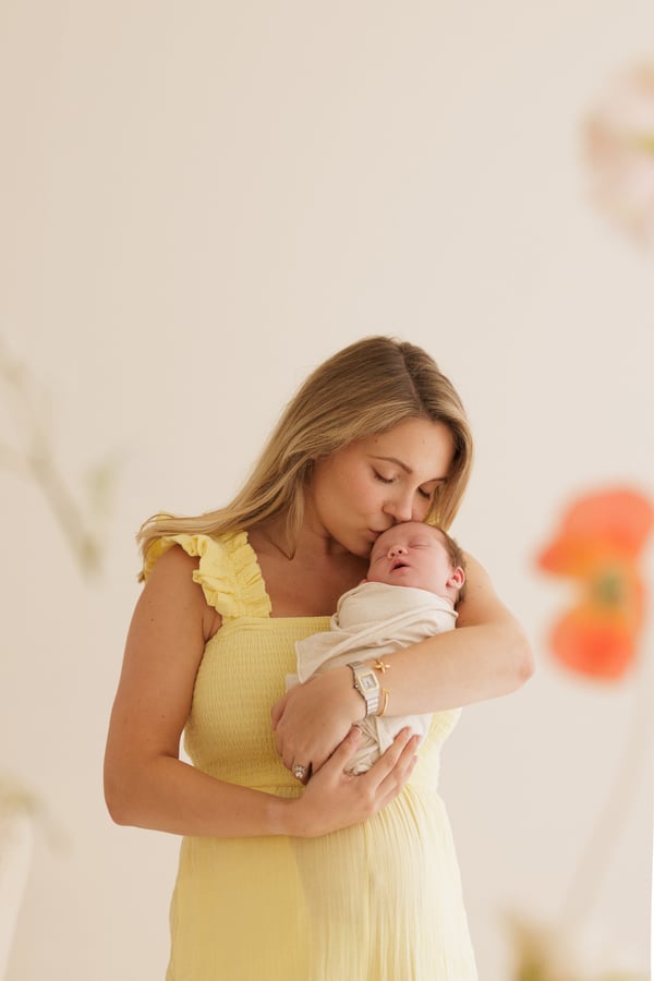 A mother in a yellow dress kissing her newborn's head, flowers out of focus behind
