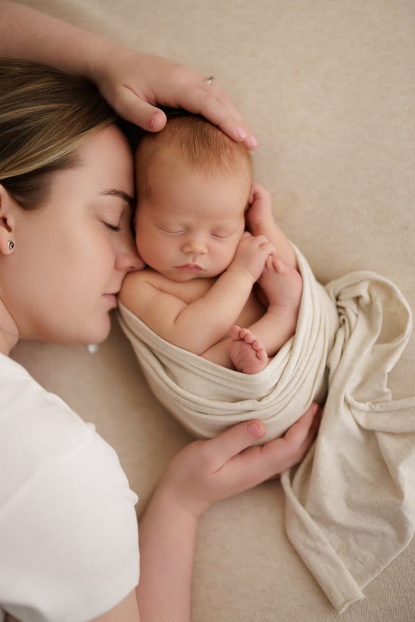 A mother lying beside her sleeping newborn, one hand resting on the baby's head