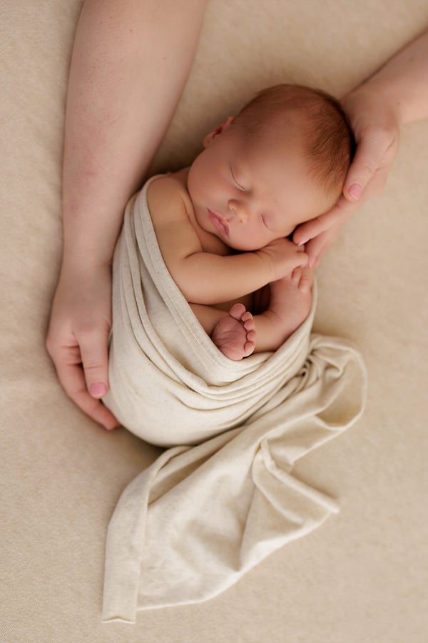 A newborn in a cream wrap, held in a mother's hands and seen from above