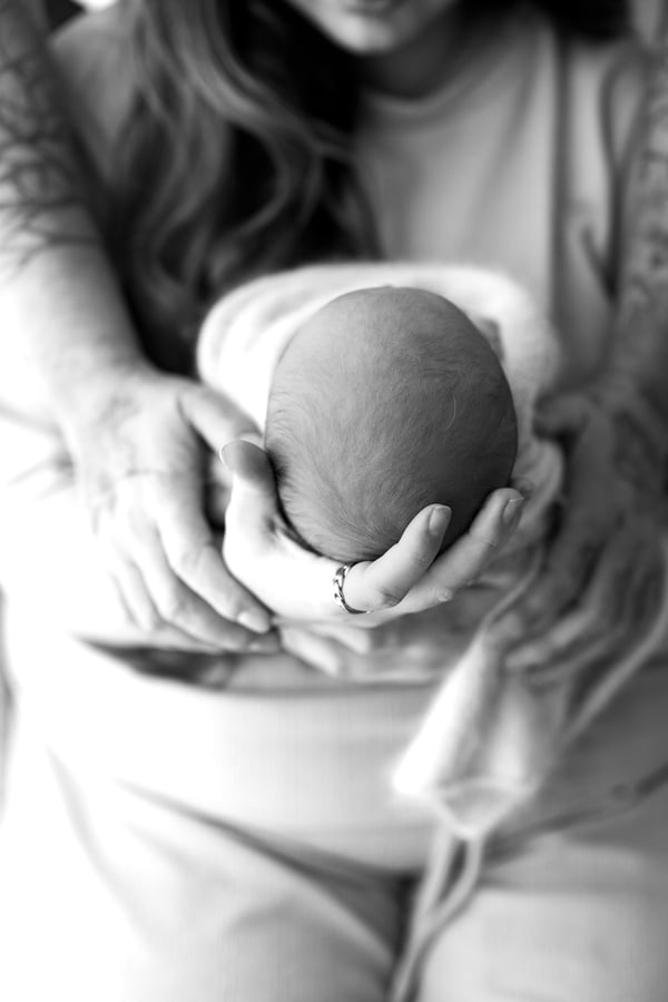 A newborn's head cradled in a parent's hands and held against their chest, the parent's tattooed arm around them