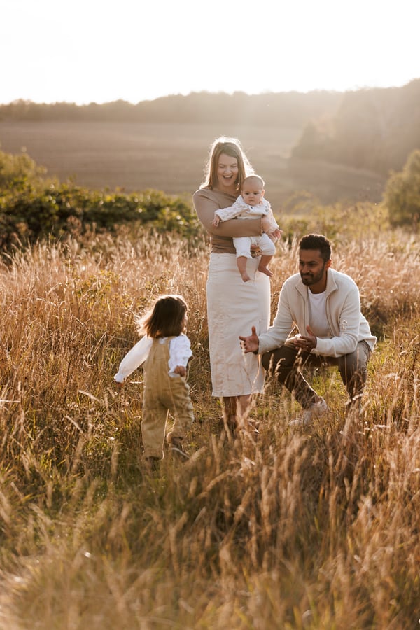 A family of four in a hillside field at golden hour, one parent holding the baby while the older child walks towards them through tall grass