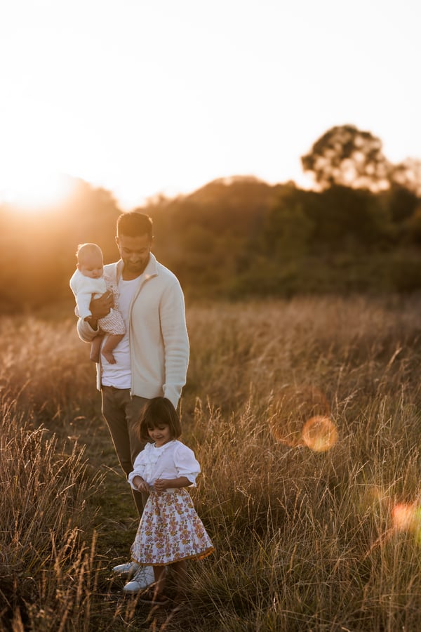 A father walking through a field carrying the baby while the older child follows through the grass, the sun low behind them