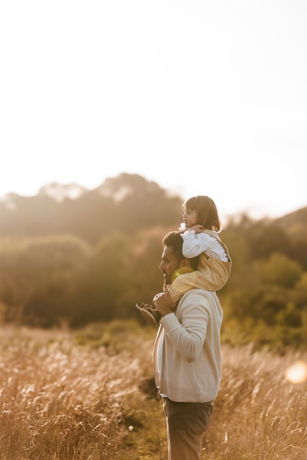 A father standing in a sunlit field with his child up on his shoulders, seen from a distance