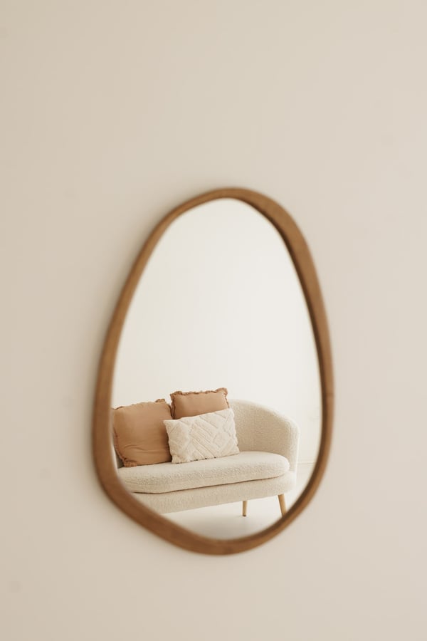 An irregular wood-framed mirror on a cream wall, with the sofa reflected in it