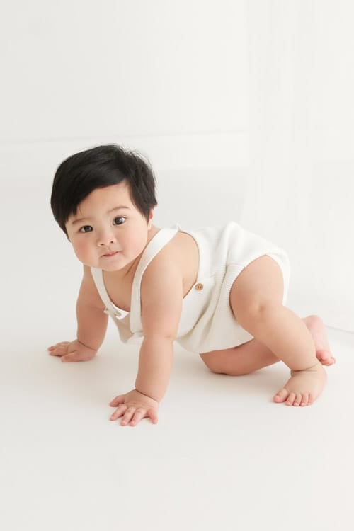 Baby crawling towards the camera on a pale backdrop, in a cream knitted romper