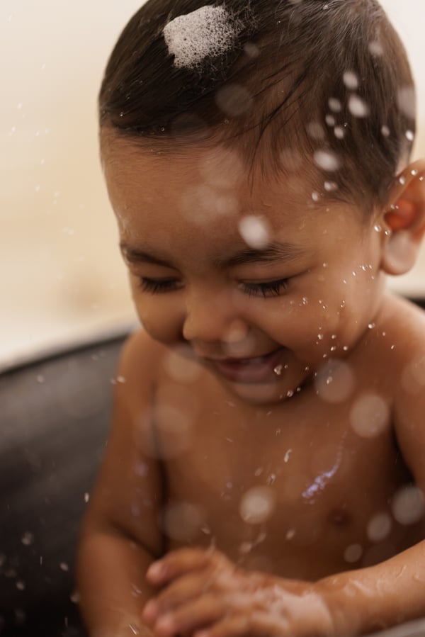 A baby laughing in the bath, water splashing around them and foam on their head
