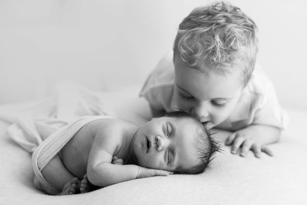 A big brother leaning over to kiss his newborn sister, in black and white