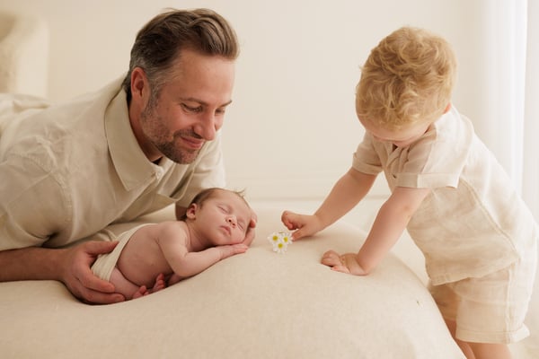 A big brother reaching out to offer a small toy to his newborn sister, his father beside them