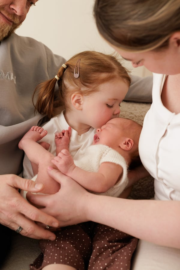 A big sister and her mother both leaning in to kiss the newborn