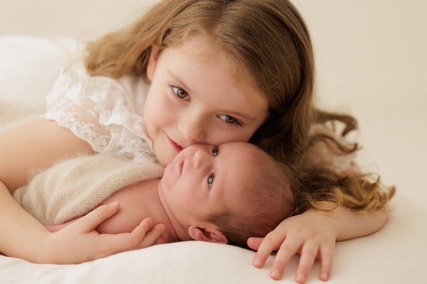 A big sister in a white lace dress lying cheek to cheek with the newborn, who is awake and looking at the camera