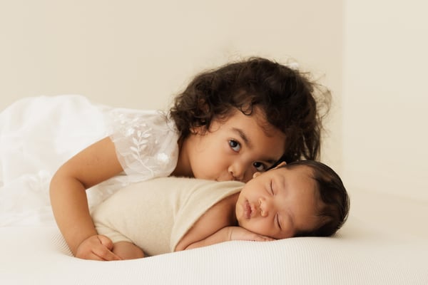 A big sister in a white dress leaning down to kiss her sleeping newborn sibling