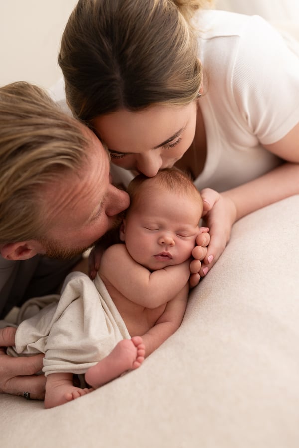 Both parents leaning in over their sleeping newborn, the father's lips against the baby's head