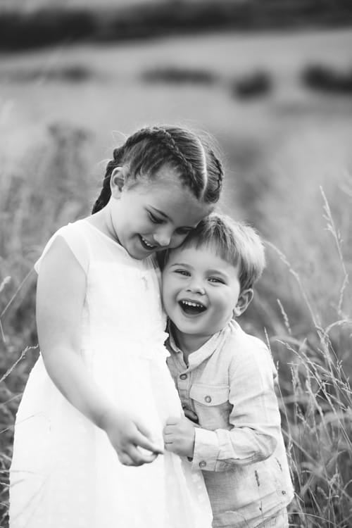 An older sister hugging her little brother in a field, both laughing