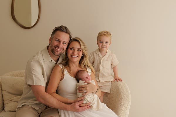 Both parents seated with the newborn while the big brother stands next to them