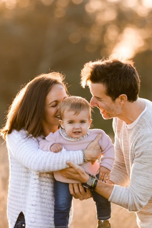 Two parents smiling down at the baby they are holding, sun behind them