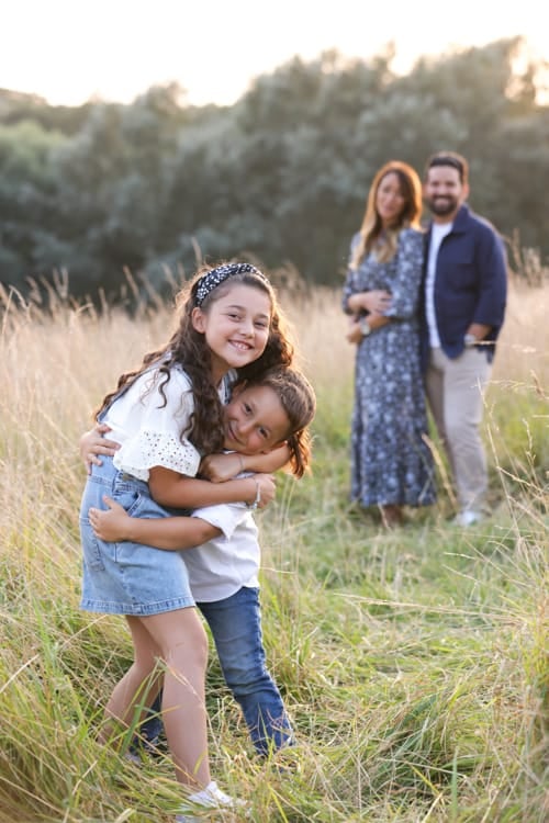 Two sisters hugging in long grass with their parents standing behind them