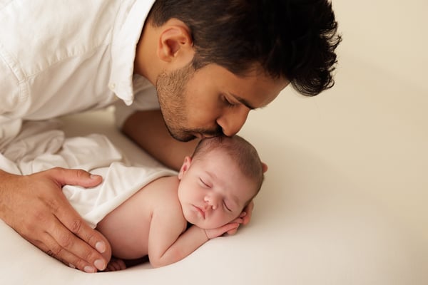 A father kissing his sleeping newborn's head, both hands cradling the baby