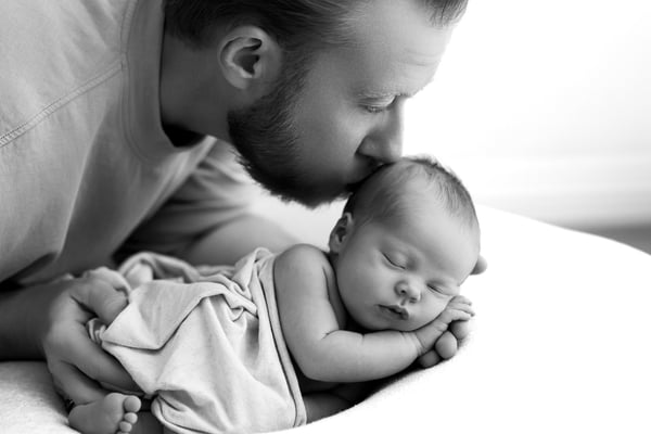 A father kissing his sleeping newborn's head against a bright white background