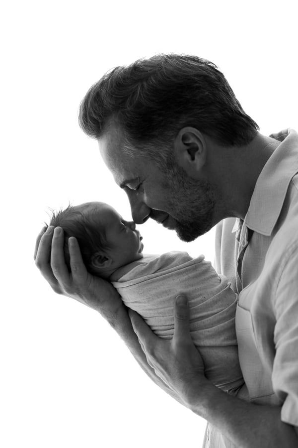 A father kissing his swaddled newborn's head against a bright window, in black and white