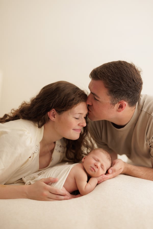 A father kissing the mother's cheek as they lie beside their sleeping newborn