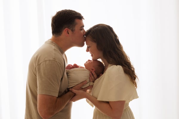 A father kissing the mother's forehead while she holds their newborn, backlit by a window