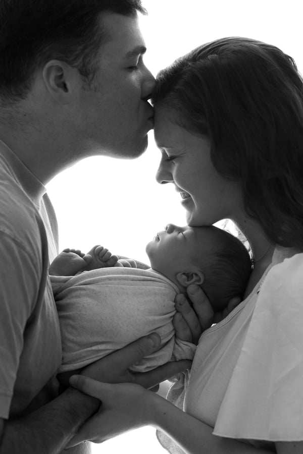 A father kissing the mother's forehead as they both hold their swaddled newborn, in black and white