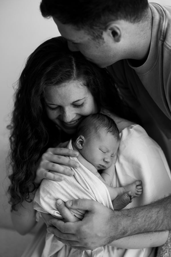 A mother holding her swaddled newborn against her chest while the father leans in and kisses her head, in black and white