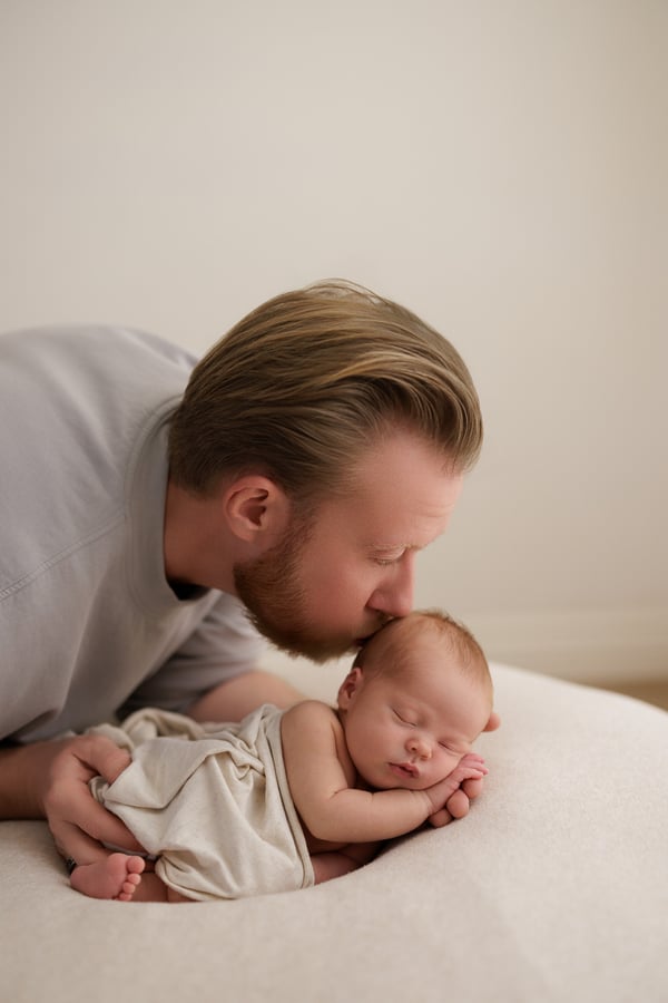 A father leaning down to kiss his sleeping newborn, who lies wrapped on a cream blanket