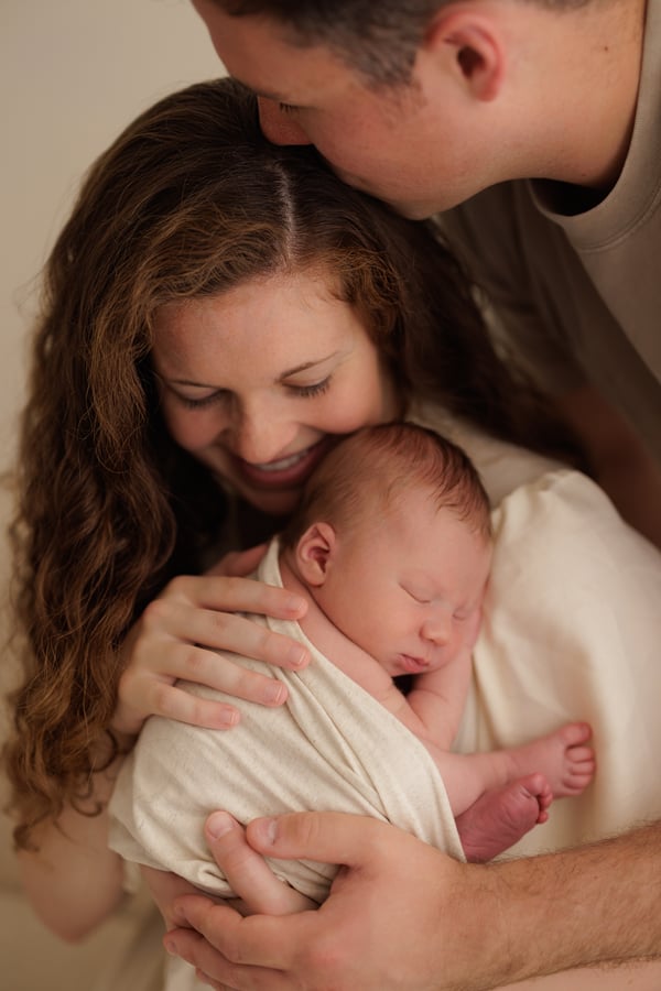 A father leaning over the mother as she holds their sleeping newborn in both arms