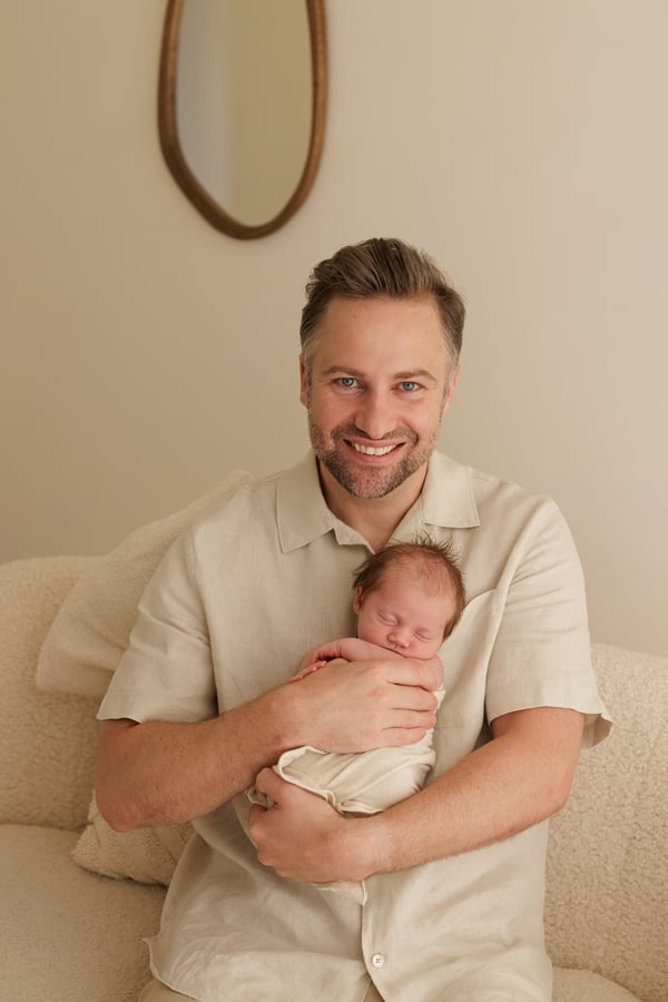 A father seated holding his swaddled newborn and smiling at the camera