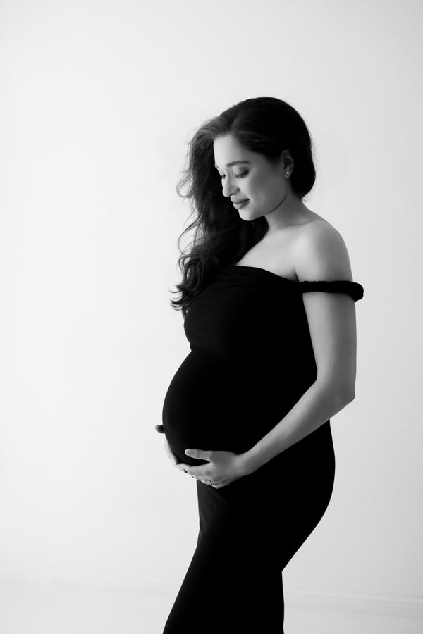 An expectant mother standing in a black off-shoulder gown with both hands on her bump, in black and white