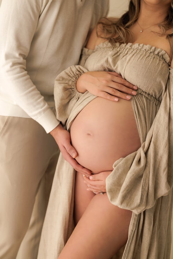 An expectant mother in a beige dress with her partner's hands resting on her bump