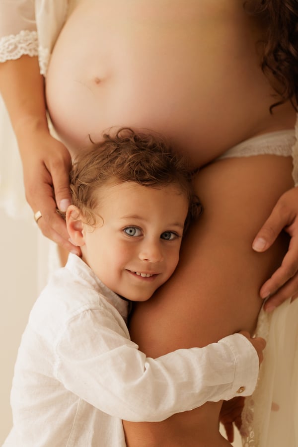 A young child hugging their mother's pregnant belly and smiling towards the camera