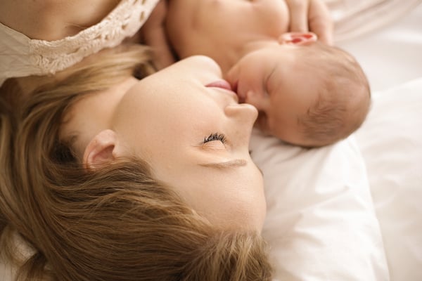 A mother lying down with her newborn beside her, their heads touching on white bedding