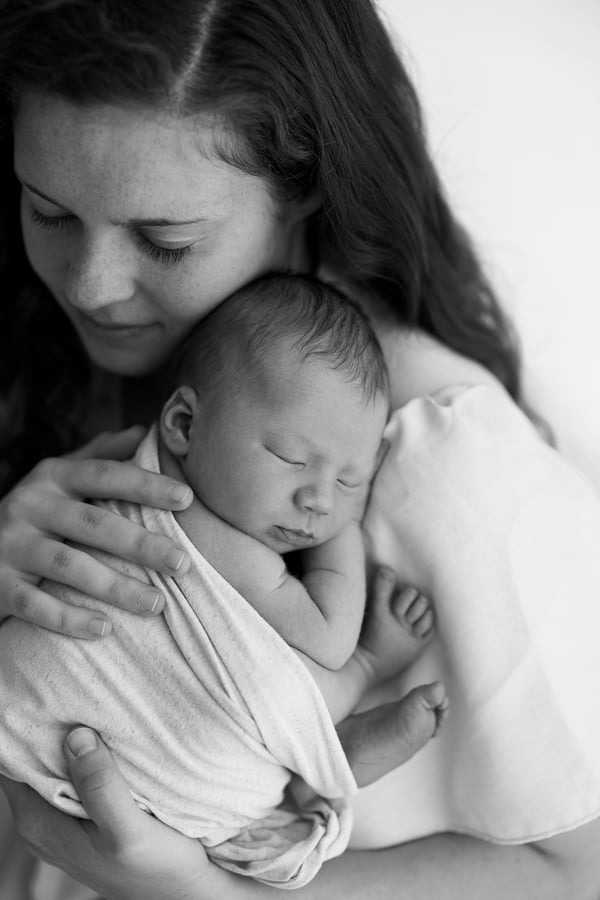 A mother holding her swaddled newborn against her chest and looking down at them, in black and white
