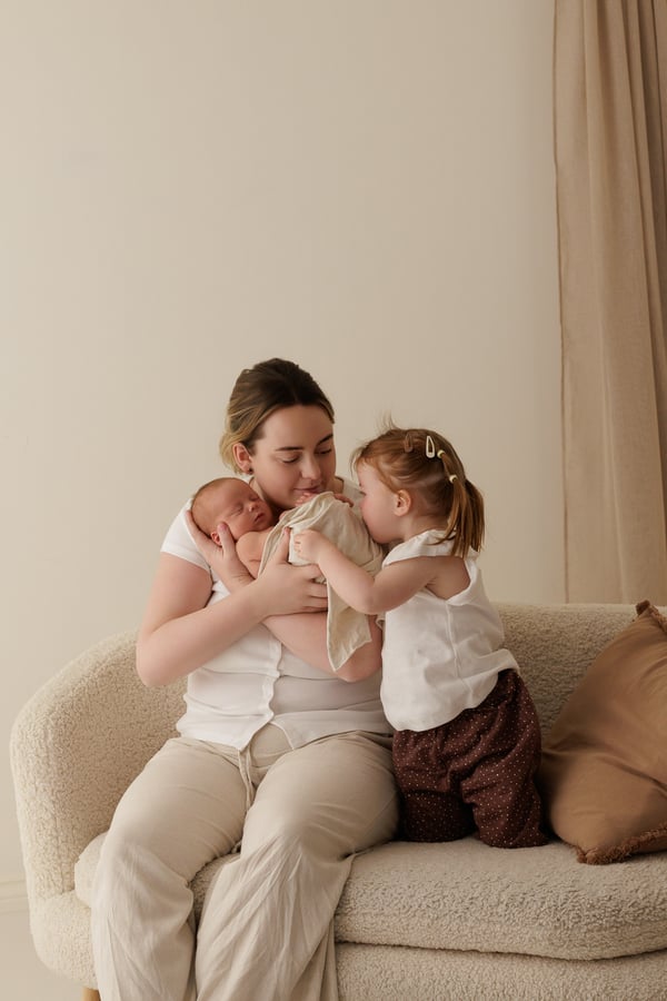 A mother sitting in an armchair holding the newborn while the big sister kneels beside them