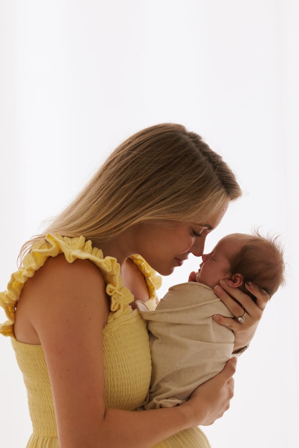 A mother in a yellow dress leaning nose to nose with her newborn against a bright window