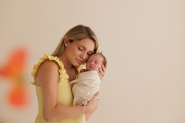 A mother in a yellow dress holding her newborn close, flowers in the foreground
