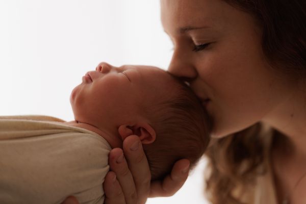 A mother holding her swaddled newborn up to her face and kissing them, against a bright background