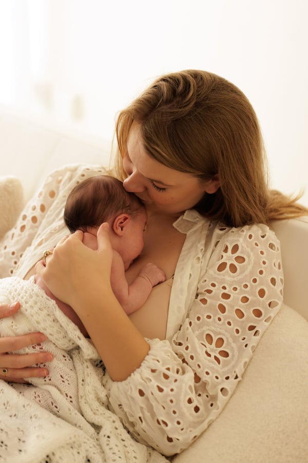 A mother kissing her newborn's head, seated beside a bright window